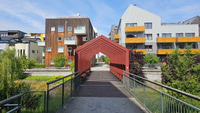 The image shows a pedestrian bridge leading to a modern residential area in Linköping, Sweden. The bridge has a red, arched canopy supported by a metal frame, and its wooden walkway is lined with metal railings. Beyond the bridge, contemporary apartment buildings with varied facades and balconies are visible. Some buildings feature dark wood paneling, while others are clad in grey and white materials with vibrant yellow balconies. Green trees and lush vegetation surround the buildings, creating a pleasant urban environment. The sky is blue with scattered clouds, indicating fair weather. In the distance, a person can be seen walking along a paved path. The overall scene depicts a well-designed, modern living space integrated with nature in a Scandinavian city.