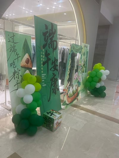 The image depicts a retail store display for the Dragon Boat Festival, situated in Liandu District, China. The foreground features clusters of green and white balloons arranged on the floor. To the left, a large green display board with Chinese calligraphy and an illustration of a zongzi (rice dumpling) is visible. Next to it, another green display board features more Chinese calligraphy and a stylized dragon boat with lotus flowers. A smaller green display board further right shows similar festive imagery. In the center, a square cardboard box with "DUAN WU" and Chinese characters is placed on the floor, adorned with illustrations related to the festival. The background shows racks of clothing within the store, illuminated by overhead lights. The floor is a polished marble finish.