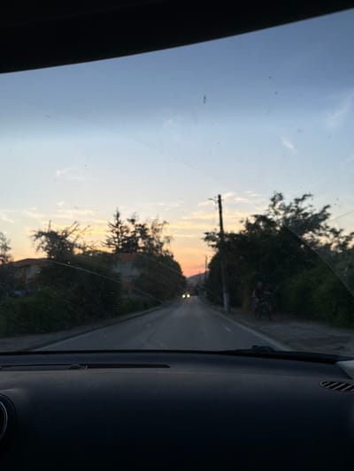 The image depicts a view from inside a vehicle, looking out through the windshield. It appears to be a sunset or dusk scene on a rural road. The sky is a gradient of pale blue to orange, with some scattered clouds. Trees and some buildings are visible along the sides of the road. A single street light is visible, and a person on a bicycle is seen in the distance. The road ahead has two sets of headlights from an oncoming vehicle. The windshield is dusty and shows some water streaks, indicating it may have recently rained or is dirty. The dashboard of the vehicle is visible at the bottom of the frame. The context provided by the query places this scene in the city of Pravets, Bulgaria.