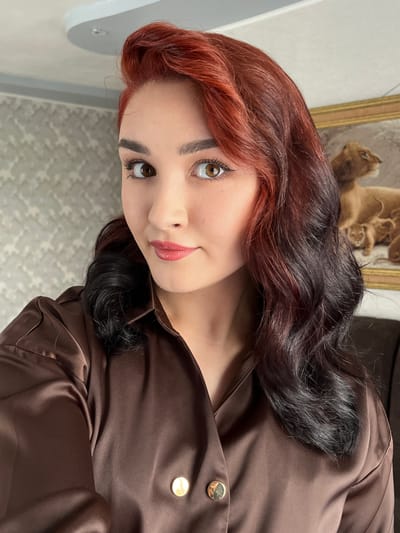 A young woman with red and black ombre hair is posing for a selfie indoors. She is wearing a brown satin shirt with gold buttons. Behind her, a painting of lions can be seen on the wall, and there is a patterned wallpaper. The ceiling has an unusual curved design with a light fixture. The image appears to have been taken in a residential setting in Starobaltachevo, Russia.