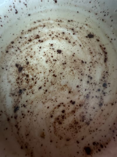 The image shows a close-up, overhead view of the surface of a beverage. The surface is covered in a light-colored foam with dark brown specks and swirls, suggesting it might be coffee with milk or a similar drink. The specks appear to be coffee grounds or chocolate powder. There are no people or identifiable objects other than the beverage itself. The location is specified as Высокая Гора, Russia, but the image provides no visual cues to confirm this or any specific urban setting.