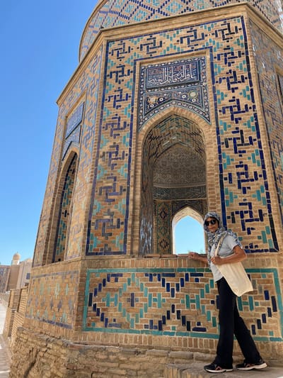 A person is leaning out of an arched opening in a building adorned with intricate blue and turquoise tilework. The building's facade features geometric patterns made of glazed bricks in shades of brown, blue, and turquoise, interspersed with brickwork. The arched opening reveals a glimpse of the sky and possibly another architectural element within. The person is wearing sunglasses, a headscarf with a colorful geometric pattern, a light-colored shirt, and dark pants. They are carrying a cream-colored tote bag over their shoulder. The scene appears to be an outdoor setting under a clear blue sky, suggesting a sunny day. The architecture and tilework are indicative of historical Islamic art and design, with the location context pointing to Samarkand, Uzbekistan, a city renowned for its rich architectural heritage. The overall impression is one of a tourist or visitor interacting with a historically significant and visually striking structure. No visible text is present in the image.
