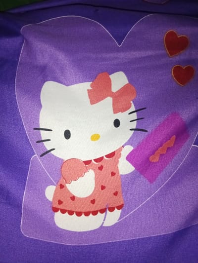 This image presents a close-up of a patterned textile, observed in the city of Ciputri, Indonesia. The fabric displays a large central graphic featuring a white stylized cartoon character, identified as Hello Kitty, enclosed within a lighter purple heart-shaped outline. The character has black oval eyes, a yellow oval nose, black whiskers, and a red bow on its left ear. It wears a light pink dress with red heart patterns, a red scalloped collar, and a red scalloped hem. The character holds a light pink, scoop-shaped object in its left hand and a purple rectangular object, possibly an envelope with a wavy reddish-orange design, in its right hand. Two smaller, solid red heart motifs are visible in the upper right quadrant, situated on the deep purple background outside the main heart outline. The dominant colors are deep purple, lighter purple, white, pink, red, yellow, and black.