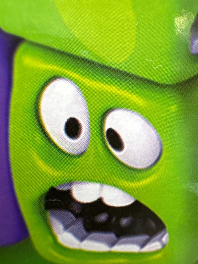 This media file is a close-up photograph of a cartoon character's face, likely from a toy or a screen. The character has bright green skin with a textured surface, resembling a fine grid pattern. Two large, oval-shaped eyes are prominent, each with a white iris and a black pupil. The eyes appear to be looking slightly upwards and to the left. To the right of the eyes is a portion of the character's mouth, which is depicted with sharp, angular teeth in black and white. The overall expression is neutral or perhaps slightly surprised. The background is predominantly green, with a hint of purple at the bottom of the frame, suggesting the character's clothing or body. The lighting is bright, casting subtle shadows that accentuate the character's features. There is no visible text in the image. The image does not provide any specific cues about the time of day, weather, or a precise location beyond the generic context of a cartoon character. The file appears to be a digital image of a printed object, possibly a sticker or a part of a larger toy.
