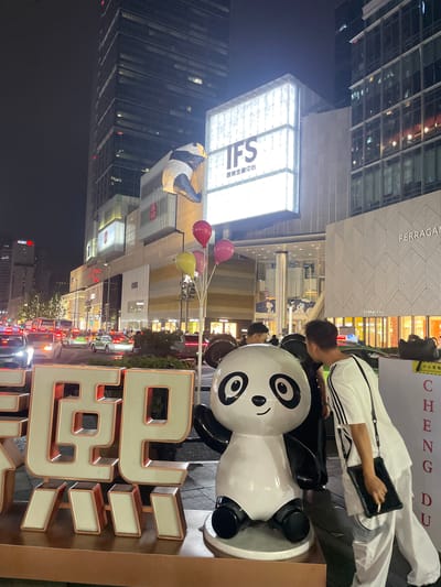 At night in Jinjiang District, China, a man in a white Adidas tracksuit, with a black bag, uses a tablet. The man is next to a large panda statue wearing a pair of black shorts. Next to the panda is a large, lighted, red-outlined sign with white characters. In the background are buildings, including the IFS building. Red, pink, and yellow balloons float above the panda statue. Cars and pedestrians are visible on the street. The Ferragamo store is also visible.