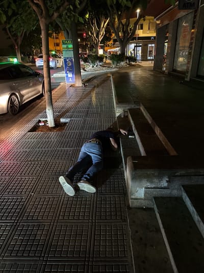 A person lies prone on a tiled sidewalk at night. The individual is wearing blue jeans, a dark shirt, and light-colored shoes. Their head is turned to the side, resting on a low concrete structure. To the left of the person, a silver car with its headlights on is parked. Further down the street, a taxi sign is visible, along with other vehicles and buildings. The scene appears to be on a city street in Eivissa, Spain, characterized by trees, streetlights, and storefronts.