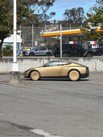 The media file depicts a side view of a futuristic, light gold car parked on an asphalt surface. The car has a sleek design with a unique, flat-topped wheel on the visible rear wheel. Its windows are tinted, and the overall aesthetic suggests a modern electric vehicle. The car is positioned in what appears to be a parking lot or roadside area.
Behind the car, a concrete wall runs horizontally, and beyond that, a chain-link fence encloses a view of several other vehicles parked in a row. These vehicles include a dark blue SUV and a silver SUV, with some hinting at a Jeep brand. A Shell gas station canopy with its signature red and yellow branding is visible in the background, suggesting the location is near a gas station. Trees with green foliage are present on the left side of the image, and a few buildings are visible on a distant hill.
The lighting indicates it is daytime, with shadows cast by the car and other objects, suggesting either late morning or afternoon. The sky is clear and blue. The presence of the gas station and the variety of vehicles imply a public or commercial setting, possibly a retail area or a service station.
There is some visible text in the image, though it's partially obscured and rotated. On a white pole in the foreground, text reads "ME" vertically, likely part of a larger word or sign. In the background, near the fence, a sign reads "END FACTORY PARKING". The overall scene conveys a sense of everyday activity in a suburban or commercial environment under clear weather conditions.