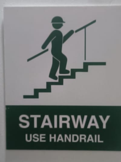 A green and white sign displays a pictogram of a person wearing a hard hat walking up stairs, holding onto a handrail. Below the pictogram, the words "STAIRWAY" and "USE HANDRAIL" are printed in white capital letters on a dark green background. This sign is located in the village of Kosshagyl in the Kosshagyl rural district of Kazakhstan. The sign is mounted on a white wall. The lighting is even, and the image is taken at a moderate distance, showing the full sign and its immediate surroundings. The image quality is clear enough to read the text and discern the details of the pictogram.