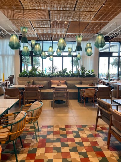 The image depicts an interior scene of a restaurant or cafe. The setting is modern and chic, with a focus on natural materials like rattan and woven textures. Hanging green pendant lights, in various sizes and shapes, are a prominent decorative element, suspended from the ceiling. The walls feature a textured, woven pattern in warm, earthy tones.
In the mid-ground, a long, built-in sofa is adorned with lush green plants, creating a biophilic design. Several rattan chairs with cushioned seats are arranged around small wooden tables. Through large windows, a glimpse of the outdoors is visible, showing trees and natural light, suggesting it's daytime. The floor is a striking mosaic of square tiles in a geometric pattern of red, green, beige, and cream.
In the foreground, two rattan chairs with light-colored cushions are placed. The overall atmosphere appears relaxed and inviting, suitable for dining or socializing. There is no visible text in the image. The location context of Ras Al-Khaimah, United Arab Emirates, implies an outdoor temperature that would allow for such an open and airy interior design.