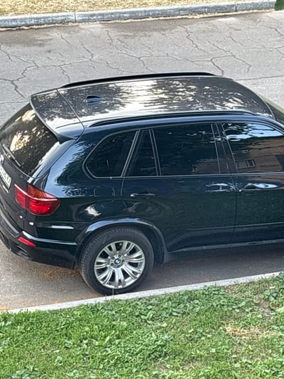 The image shows the side and rear of a black BMW X5 SUV parked on the side of a cracked asphalt road. The car's glossy finish reflects the surrounding environment, including trees and buildings. To the right of the car is a patch of green grass, bordered by a white line. The scene appears to be outdoors, with natural lighting suggesting it is daytime, likely late afternoon or early evening due to the long shadows and warm tones in the reflections. The orientation of the image is rotated, making the car appear sideways. There is some visible text on the license plate, but it is mostly obscured and illegible. The setting suggests a residential or suburban area. No people are visible in the image.