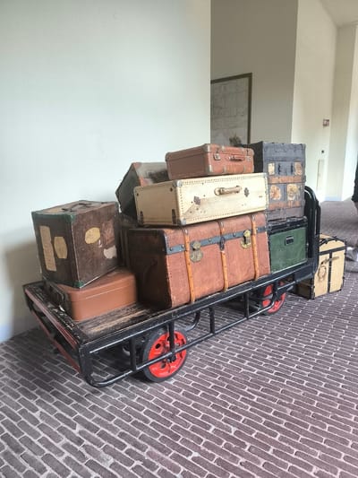 A black metal luggage cart, with a wooden platform and red wheels, is loaded with various antique suitcases and trunks. The trunks are of different sizes, colors, and materials, including brown leather, green canvas, and off-white. The cart is positioned on a brick floor, and a light-colored wall is visible in the background. A framed picture is hanging on the wall behind the cart. The scene is located inside a building, potentially in the city of Utrecht, Netherlands. A person is partially visible on the right side of the image.