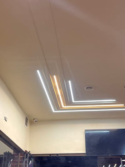The image displays an indoor environment, focusing on a light-colored ceiling and upper wall sections. The ceiling incorporates a contemporary lighting design featuring embedded linear LED strips arranged in nested L-shaped patterns. The two innermost strips emit a warm yellow light, while the two outermost strips radiate white light. Two circular ventilation grilles are visible in the ceiling on the right side.
The walls are a uniform light beige. A white, dome-shaped CCTV camera is mounted high on the left wall. A rectangular ventilation grille is positioned below and to the left of the camera. Further left on the wall, a small dark rectangular fixture is present. A large, dark-screened flat-panel television is mounted on the right wall. Below the television, a dark, reflective surface, likely a counter or dark paneling, is visible. The extreme left edge of the image shows portions of a dark structural beam and a textured reddish-brown surface, possibly brick or decorative panels, with vertical illuminated elements. No individuals are depicted in the scene. The precise location within the city of Homs, Syria, cannot be ascertained from the visual information provided.