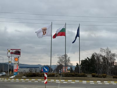 The image displays a static outdoor scene at a road intersection or roundabout in Dragushinovo, Bulgaria, under an overcast sky. In the foreground, a paved road surface with a distinctive white and yellow checkered curb line is visible. A blue circular sign with a white arrow pointing diagonally right and downwards is mounted on a red and white striped pole, positioned centrally in the lower foreground.
Three flags are prominently displayed on separate poles, flying from left to right: a white flag with a central maroon or reddish-brown emblem, the Bulgarian national flag (horizontal white, green, and red stripes), and the European Union flag (blue with twelve yellow stars in a circle).
To the left of the flags and the foreground road, a Shell gas station is present. It features a tall sign structure with a rectangular billboard displaying a red sports car and a "P" (parking) symbol. Below this, the Shell logo is visible, followed by digital price displays for various fuel types (e.g., V-Power, Diesel, A95, LPG, with values such as 2.66, 2.74, 2.50, 1.25). A blue and white gas station canopy and pumps are discernible further back. Several vehicles are parked or stationary in the vicinity of the gas station.
The central part of the intersection appears to be a planted island, featuring low green shrubs and a variety of trees, some bare deciduous and others evergreen. Distant hills or mountains are visible on the horizon beneath the grey, cloudy sky. Overhead, two parallel power lines traverse the upper portion of the frame. No individuals are present or engaged in any activity within the captured scene.