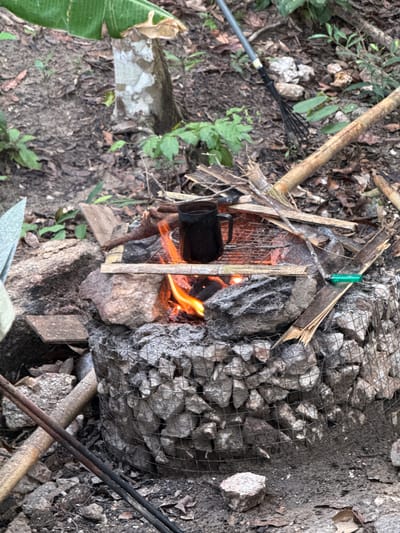 The image depicts a makeshift outdoor cooking setup in what appears to be a wooded area. A black pot is positioned over a small fire built within a structure of rocks, wire mesh, and wooden supports. Vegetation and leaf litter cover the ground around the setup. Tools or materials are scattered nearby, including bamboo poles, a rake, and potentially other implements obscured by the environment. The scene suggests a primitive cooking method. Moo 7, Thailand, is presumably the location based on provided information.