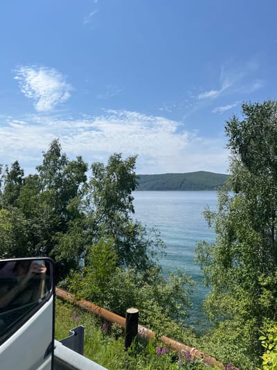 The image depicts a scenic view of a large body of water, likely a lake or sea, with a forested hill in the distance under a bright blue sky with scattered white clouds. Lush green trees and foliage frame the foreground and the water, with some rust-colored pipes and a wooden post visible near the bottom of the frame. In the lower left corner, a reflection in a vehicle's side mirror shows a person holding a phone, suggesting the photo was taken from inside a moving vehicle. The environment appears to be natural and possibly rural.