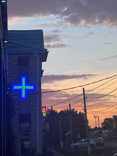 The image captures a street scene in Yegorlykskaya, Russia, during a sunset. A building with a green-tiled roof is visible on the left, featuring a prominent blue and green illuminated pharmacy cross sign. Further down the street, another illuminated cross sign with rainbow colors is partially visible. Power lines crisscross the sky, silhouetted against the colorful sunset clouds. Several vehicles are present on the road, including a light-colored hatchback and a dark sedan. Trees line the street, and streetlights and traffic signals are also discernible. The overall atmosphere is that of a calm evening in a provincial town.