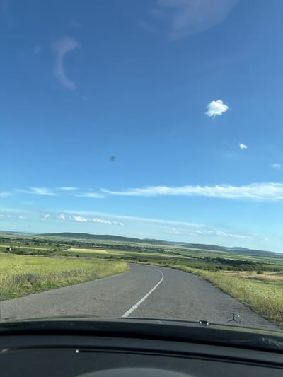 The image shows a view from inside a vehicle, possibly a car. The road is visible, turning slightly with a white line marker. The surrounding environment consists of fields of tall grass and some trees. A line of hills is visible in the background. The sky is mostly clear blue with some scattered clouds. The scene is from Kovachite, Bulgaria.