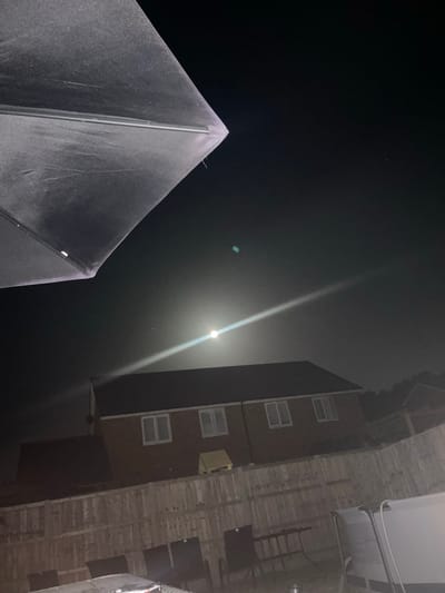The image shows a night scene with a low-angle view looking up. A geometric, pyramid-shaped object is in the lower left. A tall building with several windows is on the right. Above the building is a beam of light from the moon. The location is likely Tonbridge and Malling, United Kingdom.