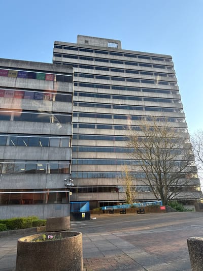 The image displays Grosvenor House, a Brutalist concrete office building located at Cumberland Place in Southampton, United Kingdom. The structure comprises a taller section of approximately 15 stories and an adjacent shorter section of about 7 stories, both characterized by exposed, textured concrete facades and horizontal bands of dark-paned windows.
On the windows of the shorter building section, multiple "TO LET" signs are visible, advertising commercial space ranging from 5,000 to 50,000 square feet, with associated branding including "BULB" and "F1-R1M". A large, bare-branched deciduous tree stands in the mid-ground on the right, partially obscuring the taller building.
The foreground features a paved plaza composed of concrete slabs and block paving. Two weathered, oval concrete planters are positioned in the immediate foreground, one containing minimal vegetation. A blue metal railing system, accompanied by several dark-colored benches, runs along the base of the buildings, suggesting a terraced area or ramp. A yellow warning sign is visible near the blue railing on the left. Two white security cameras are mounted on the exterior of the lower section of the shorter building. The sky is clear and blue, and the scene is illuminated by natural daylight. No individuals are present in the depicted scene.