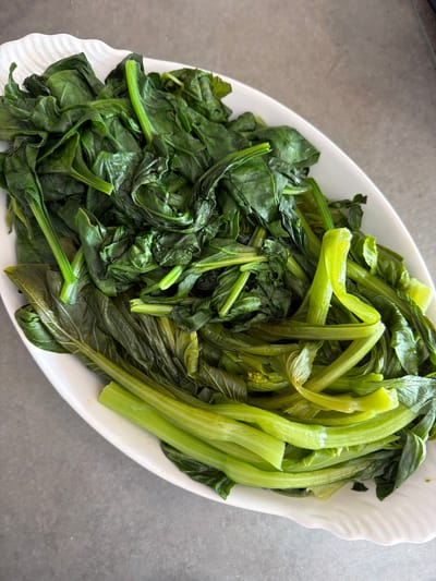 A close-up shot shows a white, oblong serving dish filled with cooked leafy green vegetables. The vegetables appear to be divided into two main sections. One section consists of smaller, darker green leaves, seemingly cooked spinach or a similar leafy green. The other section features lighter green, thicker stalks and leaves, possibly Bok Choy or Choy Sum. The dish is set against a smooth, gray surface. The image does not depict any elements indicating a location in Champs-sur-Marne, France, or any people, actions, or interactions.