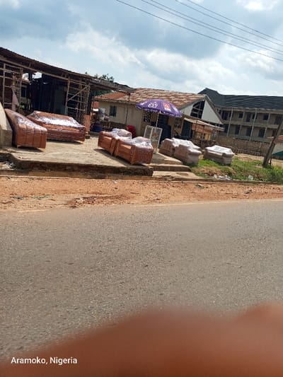 The image shows a street scene in Aramoko, Nigeria, located at latitude 7.827 and longitude 4.518. Several brown leather couches are lined up in front of a small shop with a purple umbrella. The shop is in the middle of the frame, with a two-story building behind it on the right side. The street is paved and empty. The sky is partly cloudy. The couches are likely for sale. The image was taken from a low angle, looking down at the scene.