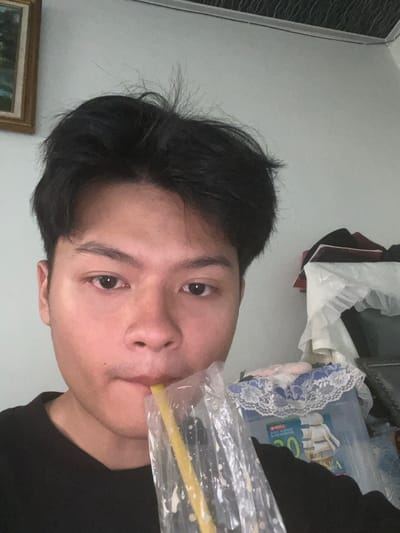 The image shows a close-up, rotated view of a young person with dark hair, looking into the camera. They are wearing a black shirt. The person is holding a clear plastic bag filled with a white, frothy substance, from which a yellow straw extends. The person's mouth is around the straw, suggesting they are drinking from the bag.
The background is a plain, light-colored wall. To the left, a framed picture is partially visible. To the right, out of focus, is a cluttered area with various items, including what appears to be a plastic bin containing fabrics and a book with visible text. Some of the text is in Indonesian, including the word "BUANA" which is clearly legible on the book cover.
The lighting suggests it is indoors, possibly during the day given the bright, even illumination. There are no explicit cues about the weather or specific time of day, other than it being indoors. The person's expression is neutral. The scene appears to be a casual, personal setting.