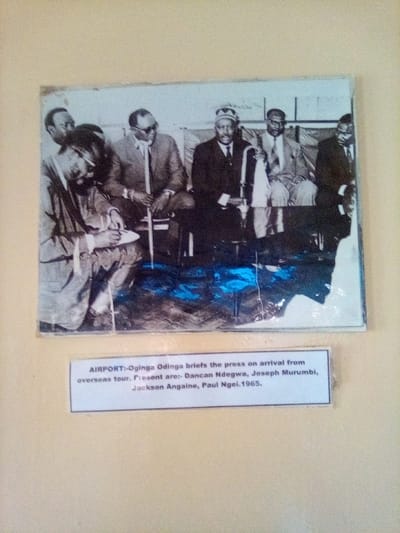 The image displays a photograph of an aged, faded, black-and-white picture mounted on a light beige wall. The depicted scene, identified by an accompanying caption as an airport press briefing from 1965, features approximately seven to eight African men.
In the foreground of the black-and-white photograph, a man with a beard and dark glasses is seated on the far left, appearing to take notes on a white pad. To his right, five other men are seated in a row, dressed in suits. One man on the left of this seated group wears dark glasses and holds a cane. Another prominent figure among the seated men wears a dark suit and a distinctive patterned cap or headwear. Other men in suits are visible, some partially obscured.
A white caption label is affixed directly below the photograph, stating: "AIRPORT:-Oginga Odinga briefs the press on arrival from overseas tour. Present are:- Dancan Ndegwa, Joseph Murumbi, Jackson Angaine, Paul Ngel.1965." This text identifies the primary subject as Oginga Odinga briefing the press, with Dancan Ndegwa, Joseph Murumbi, Jackson Angaine, and Paul Ngel among those present, in 1965 at an airport.
The historical photograph exhibits significant wear, including faded tones, visible creases or damage, and a prominent blue discoloration or reflection spanning the lower section. The overall presentation indicates a historical artifact displayed for public viewing. This entire displayed image is physically located in the city of South Sakwa ward, Kenya.