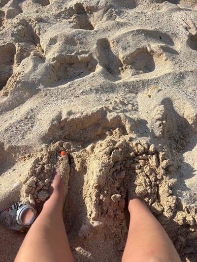 An overhead view depicts two human legs partially submerged in light brown sand at a beach, identified as Olde Yard Village, British Virgin Islands. The legs are bare, appearing tanned, and extend from the bottom of the frame towards the upper center. The left leg has a small, orange, spherical object resting on the sand near its toes. To the immediate left of the left leg, a crumpled, silver-colored aluminum beverage can is visible, resting on the sand. The sand itself is textured with various mounds, depressions, and scattered small particulate matter. Shadows are cast across the sand from the objects and undulations, indicating direct sunlight from the upper right. In the upper right quadrant of the frame, several small, pale yellow, curved objects are partially embedded in the sand. No other individuals or significant actions are depicted.