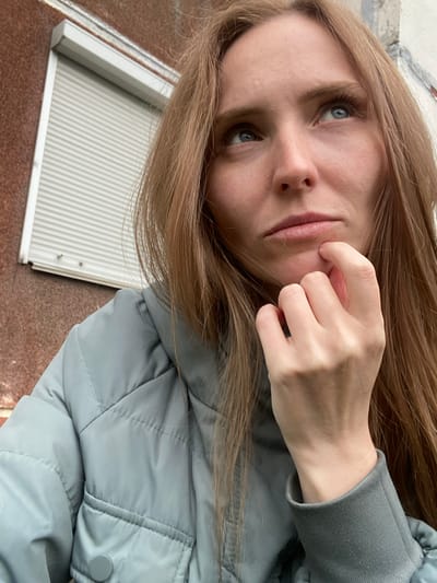 A young Caucasian woman with long, straight, light brown hair is captured in a close-up, waist-up shot, looking upwards and to the right. She is wearing a light blue puffer jacket over a gray sweatshirt. Her right hand is resting on her chin, with her index finger touching her lower lip, suggesting a pensive or contemplative mood. The background is out of focus, showing a textured, reddish-brown wall on the left and a portion of a building with a light-colored, ribbed shutter on the right. The overall lighting appears to be overcast. The image was taken in the city of Yuzhno-Sakhalinsk, Russia.