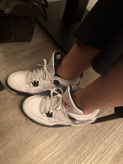 A person is sitting in a chair, their legs are bent, and they are wearing white Air Jordan 4 sneakers with black accents and white laces. The sneakers are slightly scuffed, and the laces are tied. The background is a light-colored wooden floor, and part of a dark-colored bag is visible to the left. The person is wearing dark-colored clothing.