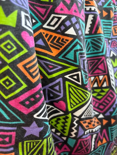 The image is a close-up, high-angle shot of a black fabric with a vibrant, geometric pattern. The pattern consists of various shapes, including triangles, squares, diamonds, and zigzags, rendered in bright neon colors such as pink, lime green, orange, purple, and turquoise. The fabric appears to be folded or draped, creating shadows and highlights that accentuate its texture and the folds of the material. There are no people or discernible environmental elements present, suggesting the image focuses solely on the textile design. The image does not provide any information about its location in Blagoveshchensk, Russia.