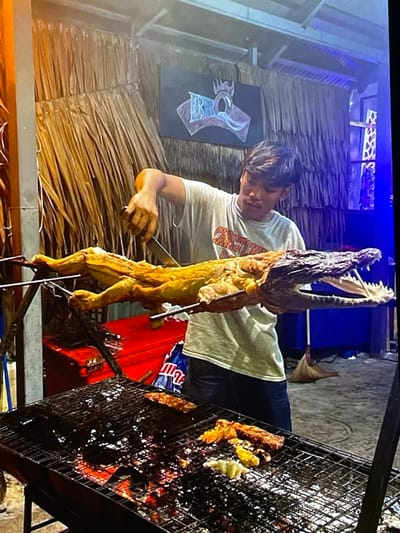 A man is roasting a whole alligator on a spit over a charcoal grill. The alligator is golden brown and appears to be cooked. The man is wearing a white t-shirt and dark pants. He is holding a knife and fork, preparing to carve the alligator. The grill is covered in grease and char, with a few pieces of meat cooking on it. In the background, there is a thatched-roof structure and a sign with the word "BBQ" on it. The scene appears to be an outdoor market or restaurant in Harmanli, Bulgaria.