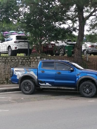 In the city of Xuân Hương - Đà Lạt, Vietnam, a blue Ford Ranger Raptor pickup truck is parked on the side of a road. The truck has a black roll-up tonneau cover and "RAPTOR" decals on its side. In the background, a white SUV is parked behind a metal fence, with several colorful umbrellas visible in the distance. A large tree with thick branches stands to the right of the truck, partially obscuring the view of other vehicles and a green trash bin. The scene appears to be in a public area, possibly near a park or tourist attraction.