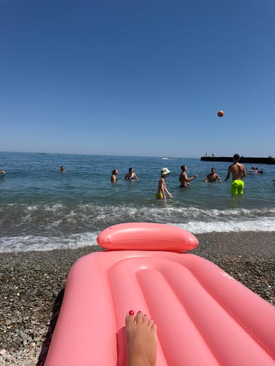 This image depicts a beach scene on a clear, sunny day, with the Black Sea providing a backdrop. In the foreground, a pink inflatable mattress is partially visible, with a person's foot, sporting red nail polish, resting on it. The beach itself is composed of pebbles.
In the water, several individuals are engaged in various activities. Some are swimming, while a group appears to be playing with a ball. The water is calm with gentle waves lapping at the shore. In the distance, a pier or breakwater is visible, with a few people standing on it. There are also some boats or watercraft further out in the sea.
The sky is a vibrant blue, with no clouds present. The overall atmosphere is one of leisure and recreation. The location is identified as a beach in the city of Partenit, Ukraine.