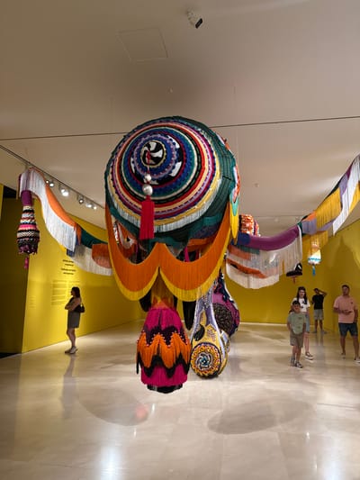 The image depicts a large, colorful, crocheted textile sculpture hanging from the ceiling in a museum gallery. The sculpture is predominantly made of concentric circles of various colors, with intricate patterns and many hanging fringes and tassels. It appears to be suspended by thin wires. In the background, a bright yellow wall features text in both Spanish and English, and a woman in a gray dress stands looking at the artwork, with her arms crossed. Several other people, including children, are visible in the middle ground, observing the sculpture. The floor is polished marble, reflecting the artwork and the people. The lighting in the gallery is primarily from ceiling-mounted spotlights. The scene is consistent with an exhibition at the Centro Pompidou Málaga, given the distinctive yellow exterior visible in other views of this location and the modern art installations often found there.