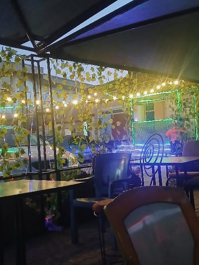 This nighttime image depicts an unoccupied outdoor seating area, likely a restaurant or cafe, located in Kwekwe, Zimbabwe. The scene is illuminated by multiple strings of warm white incandescent bulbs and additional green LED-like lights, entwined with artificial green foliage resembling vines. A dark canopy or awning covers the upper portion of the frame. Furniture includes several dark-colored tables and chairs; one chair in the immediate foreground features a light seat and an orange-brown back, while others in the mid-ground are dark metal, some with distinctive spiral backrests. In the background, behind a metal railing, a light-colored building with windows is visible, alongside a partially obscured light-colored vehicle. Ambient blue and green lights are present in the deeper background, suggesting an adjacent street or public space. No individuals are present.