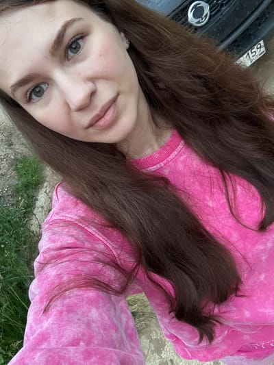 This is a self-portrait of a young woman with long, dark brown hair, taken outdoors. She is wearing a pink tie-dye sweatshirt. Her face is partially obscured by her hair and the angle of the photo, but her eyes, nose, and lips are visible. She has light skin with a rosy blush and light brown eyebrows. Her eyes are green. The background shows a surface that appears to be concrete or dirt, with some green grass and foliage. To the left, a portion of a black car is visible, including a license plate with the numbers "159" and the letters "RUS". The lighting suggests it is daytime, likely outdoors in natural light, with no indication of specific weather conditions. The overall mood appears casual and relaxed. The setting suggests a location in Nizhnyaya Gar', Russia, as indicated by the license plate.