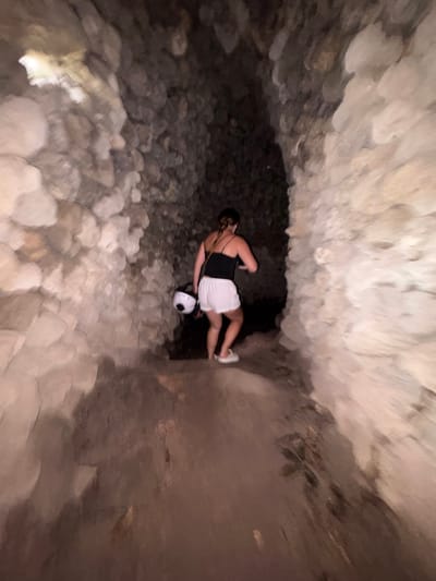 An individual is depicted exploring a narrow, rocky cave passage in Cam Ranh, Vietnam. The scene is dimly lit, typical of a subterranean environment. The person, viewed from behind and slightly to their left, is wearing a black sleeveless top, white shorts, and white sneakers. They are holding a white helmet or a spherical object with an integrated light source, which casts a bright beam onto the uneven, stone-covered cave floor directly in front of them.
The cave walls and ceiling are composed of rough, light-colored rock formations, creating a rugged and enclosed space. The ground is similarly covered with an accumulation of irregular rocks and debris, suggesting challenging terrain for traversal. The person's posture indicates they are carefully navigating or pausing during their exploration. There are no discernible external light sources, and the time of day is irrelevant within the confined cave. No text is visible within the image.