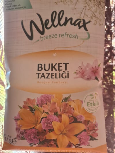 This media file is a close-up of a product packaging. The packaging features a floral design with large orange lilies and smaller pink flowers. Prominently displayed is the brand name "Wellmax" in large, cursive black lettering, accompanied by the text "breeze refresh". Below this, in vertical orientation, is "BUKET TAZELİĞİ" and "Bouquet Freshness". To the left of the floral arrangement, there is a green circle with Turkish text which translates to "Long-lasting Effective Scents". On the bottom left, there is a list of suggested uses in Turkish: "Çekmece" (Drawer), "Banyo" (Bathroom), and "Yatak Odası vb. kullanım alanları için" (for areas like bedrooms, etc.). At the bottom right, there are product specifications: "0.71 fl oz" and "21 ge". The packaging appears to be for an air freshener or a similar home fragrance product. The background is a textured, sparkly fabric, possibly a tablecloth or a decorative surface, suggesting the product is displayed for promotional purposes. There is no indication of people or a specific time of day or weather. The text is in both Turkish and English.