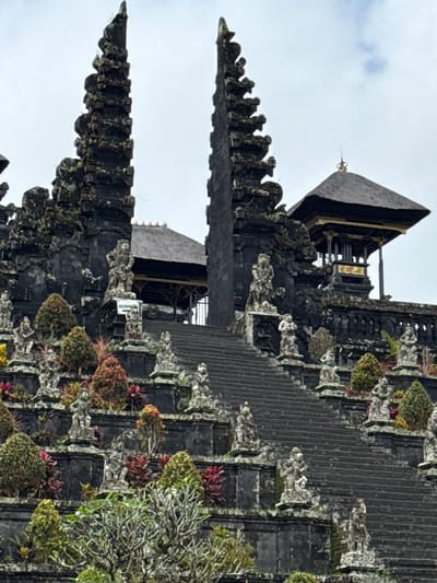 This is a vertical, eye-level, medium shot of a stone temple complex. The temple is made of dark stone and features multiple tiered structures and ornate carvings. Two tall, slender towers with intricate details flank a central, wider structure topped with a multi-tiered roof. A grand stone staircase ascends from the foreground towards the upper levels of the temple. Numerous small, stone statues of human figures are placed along the staircase and on the various platforms and ledges of the temple. Lush green and red foliage is interspersed among the stone structures and statues. The sky above is overcast with a few wisps of clouds. The location is identified as the Besakih Temple complex, situated on the slopes of Mount Agung near the village of Rendang, Bali, Indonesia. There are no people visible in the image.