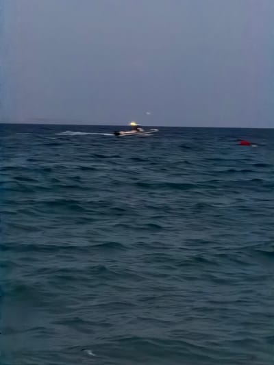 The image depicts a body of water, likely the sea, under a twilight sky. The water is dark blue with visible ripples. In the distance, a motorboat with lights on is moving from left to right, leaving a wake. On the right side of the frame, a red buoy is floating on the surface of the water. The horizon is visible as a faint line separating the water from the sky. The overall mood is calm and serene. The scene is set in the waters off the coast of Calvià, Spain, a popular tourist destination known for its beautiful beaches and coastal areas.