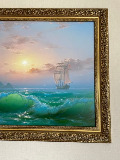 The image displays a framed oil painting of a seascape. The painting depicts a large sailing ship with multiple sails navigating through a rough sea under a dramatic sunset sky. The sun is low on the horizon, casting a golden glow and illuminating the clouds with hues of orange and pink. The sea is a vibrant turquoise, with large, cresting waves detailed with white foam. In the distance, silhouetted islands or landmasses can be seen on the left. The frame is ornate and gilded, with intricate scrollwork and decorative patterns. The painting is hung on a textured off-white wall. There is no information provided in the image or crops that links this painting to the city of Yevpatoriya, Ukraine.