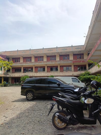 In the city of Bireuen, Indonesia, a multi-story building with beige and pale orange walls stands under a cloudy sky. The building features multiple rows of windows with dark red shutters and decorative elements along the top. In the foreground, a gravel lot is occupied by several parked vehicles. A black SUV is parked prominently, followed by a white van, and then two black motorcycles. Green foliage from trees and bushes is visible around the building and to the right of the vehicles. The scene appears to be an outdoor area adjacent to the building, possibly a parking lot or courtyard.