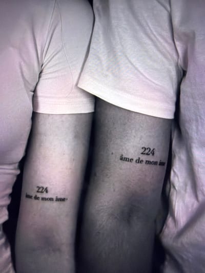 The image displays a close-up view of a person's legs, covered in white fabric from the waist down. Two tattoos are visible on the skin, one on each leg, positioned vertically. Both tattoos consist of the number "224" followed by the French phrase "âme de mon âme". The setting appears to be indoors, with soft lighting that creates a somewhat muted and intimate atmosphere. The fabric suggests comfortable clothing, perhaps pajamas or loungewear, and the overall impression is one of personal reflection or intimacy. The tattoos are the most prominent and notable details in the image.