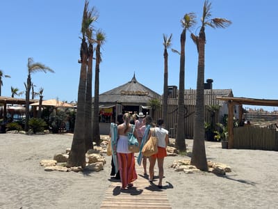 The image depicts a beach scene in the city of San Roque, Spain. In the foreground, a wooden pathway is laid out on the sandy beach, surrounded by palm trees and rocks. Four individuals, likely women, are walking away from the viewer on the pathway, heading towards a beach establishment. They are carrying beach bags and towels.
The establishment in the background appears to be a beach bar or restaurant, with a thatched roof and signage that reads "CHAMBAO". Palm trees are a prominent feature throughout the scene, their fronds rustling in the breeze against a clear blue sky. The environment suggests a sunny day at a popular beach destination.