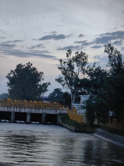 The image shows a concrete structure with an open channel of water flowing in the foreground. Above the water, there is a bridge with yellow railings and several dark rectangular openings that appear to be floodgates or culverts, with water visibly exiting from some of them. Trees are visible on either side of the structure, and a white building with a balcony is situated behind the bridge. The sky is overcast with a few scattered clouds, and the overall lighting suggests it is either early morning or late evening. The image appears to be taken from a slight elevation, possibly from a boat or another bridge, looking towards the concrete structure and the surrounding vegetation. The text "SLEEPER" is faintly visible in the sky, likely a reflection or overlay. The location is identified as Uchana, India.