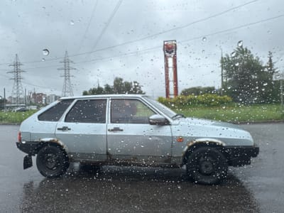 A light-colored Lada hatchback, appearing weathered and with rust spots, is parked on a wet asphalt surface, possibly a roadside, under an overcast, rainy sky. The car is positioned in the mid-ground of the image, viewed from its side. Raindrops are visible on the foreground, suggesting the photograph was taken from inside a vehicle or through a window. In the background, power lines and electrical towers stretch across the frame. Further in the distance, there are buildings, trees, and a red and yellow tower with a circular sign at the top, potentially indicating a commercial establishment. The overall atmosphere is somber, consistent with a rainy day in an industrial or semi-urban area. The presence of a Lada, a common car model in Russia, along with the specific type of power line structures and the general landscape, is consistent with a location in Tolyatti, Russia, known for its automotive industry and industrial infrastructure.