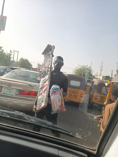 A street vendor stands between stopped cars, selling goods. He carries a large, rectangular display board loaded with items on his shoulder. In his other hand, he holds a plastic bag filled with colorful merchandise. In the background, there are yellow, three-wheeled vehicles, cars and a street sign. The perspective is from inside a car, through the windshield.