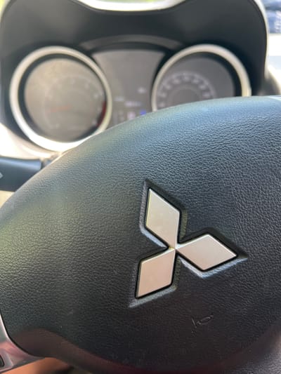 This media file is a close-up, eye-level shot of the steering wheel of a car, focusing on the Mitsubishi logo. The steering wheel is covered in black leather, with the silver Mitsubishi emblem prominently displayed in the center. The emblem consists of three diamond shapes arranged in a fan-like pattern. To the right of the emblem, a small horn icon is subtly visible on the leather.
In the background, out of focus, are parts of the car's dashboard, including two circular instrument clusters. The cluster on the left appears to be a speedometer or tachometer with a needle pointing slightly past the center. The cluster on the right is less distinct but also shows circular markings and a needle. The overall setting is the interior of a car.
The lighting suggests it is daytime, with natural light illuminating the scene. There are no people visible, and no specific activity or event is taking place beyond the implied context of being inside a parked or operating vehicle. The location is unknown, and there are no discernible weather cues. No text is clearly visible in the image, other than the horn icon.
