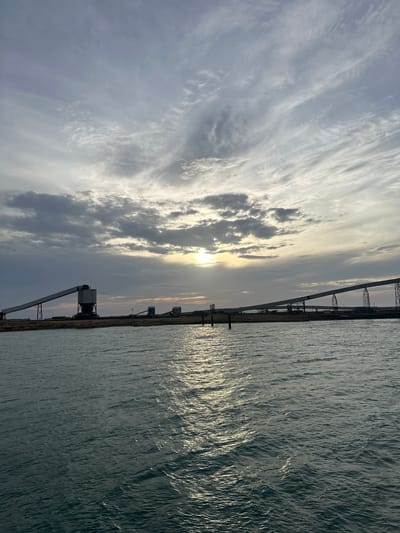 The image captures an industrial waterfront scene in Uribia, Colombia, under an overcast sky during either sunrise or sunset. The sky, occupying the upper half, displays a mixture of grey and white clouds, with a hazy sun visible slightly right of center, emitting a soft, diffused yellow light. Directly below the horizon, a low-lying landmass features multiple large-scale industrial structures. On the left, a prominent, dark-colored conveyor belt system descends at an angle towards the water, supported by metal framework and connected to a tall, box-like building or silo. To the right, additional conveyor belts extend horizontally across the land, supported by a series of metal trestles. Piles of bulk material are discernible on the land adjacent to these structures. The lower half of the image is dominated by a body of water, exhibiting small, choppy ripples. A distinct shimmering reflection of the sun's light stretches across the water from the horizon towards the foreground. No human figures or active machinery are visible.